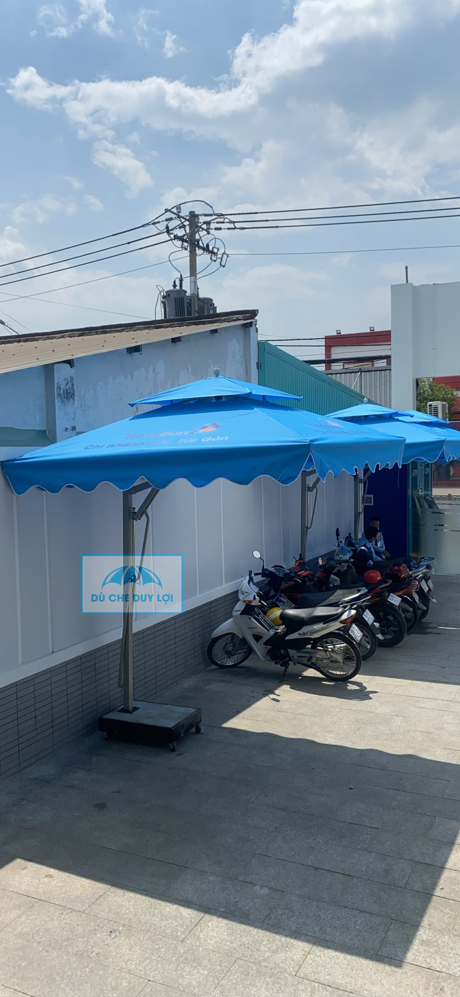 Dãy dù màu xanh che khu vực để xe, với trụ và bộ đế bố trí sát một phía.